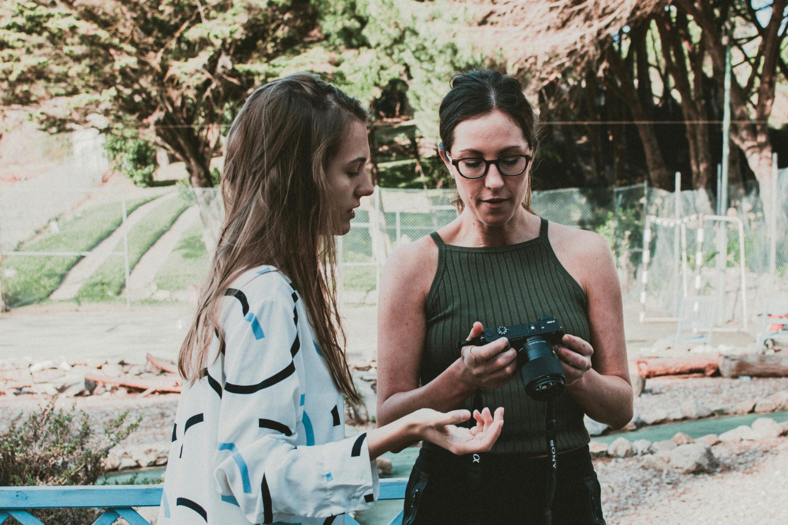 Two women in a park discussing camera settings, showcasing a casual photography lesson.